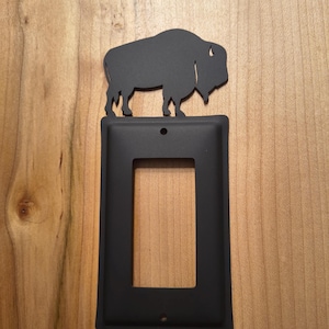 May include: A matte black metal light switch cover plate featuring a silhouette of a standing American bison attached to the top edge. The rectangular plate has a central vertical rectangular cutout and two small circular screw holes for mounting.