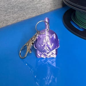 May include: A metallic purple 3D-printed keychain featuring a stylized pug head with a flat face and pointed ears. The keychain includes a silver-toned metal split ring and a lobster claw clasp, positioned next to a stack of three filament spools.