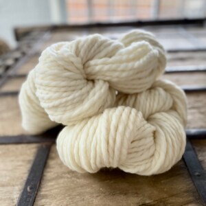 Könnte beinhalten: Eine Knäuel cremefarbenes Garn aus Naturfasern liegt auf einer Holzoberfläche. Das Garn ist weich und flauschig und eignet sich perfekt zum Stricken oder Häkeln.