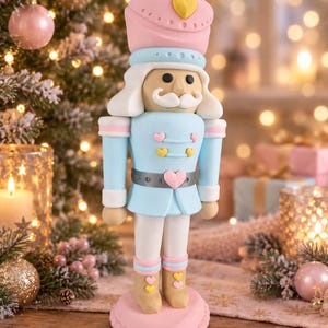 Könnte beinhalten: Eine Nussknacker-Figur mit hellblauer Jacke, weißer Hose und rosa Hut. Das Design umfasst kleine gelbe und rosa Herzakzente auf der Brust, dem Gürtel und den Stiefeln. Die Figur steht auf einem rosa herzförmigen Sockel mit weißem Schnurrbart.