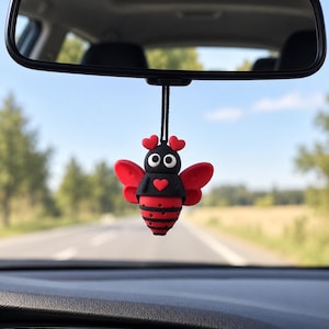 Könnte beinhalten: Ein hängendes Auto-Ornament in Form einer Biene mit einem schwarz-roten Körper, roten Flügeln, zwei herzförmigen Fühlern und einem kleinen roten Herz auf der Brust. Die Biene hat große weiße Augen mit schwarzen Pupillen und hängt an einer schwarzen Schnur.