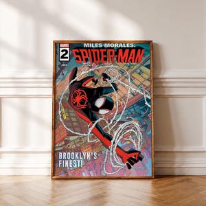 Op de afbeelding: Een ingelijste stripboekcoverprint getiteld Miles Morales: Spider-Man nummer 2. Het kunstwerk toont het personage in een zwart en rood pak dat door een stad slingert met witte webben. De tekst luidt Miles Morales: Spider-Man en Brooklyn's Finest.