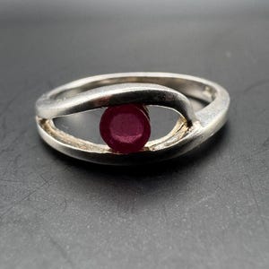 Op de afbeelding: Een zilveren ring met een gespleten schachtontwerp dat rond een enkele, ronde, dieprode edelsteen in het midden buigt. De metalen band heeft een gladde, gepolijste afwerking en de steen is geplaatst tussen de twee open metalen armen.