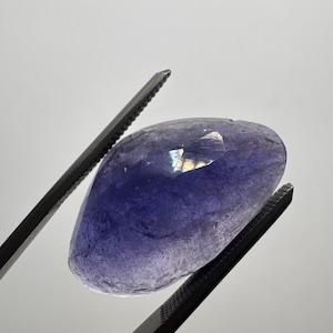 May include: A faceted, translucent purple-blue gemstone held by a pair of metal tweezers. The stone features a polished, irregular shape with visible internal inclusions and light reflecting off its top surface against a plain background.