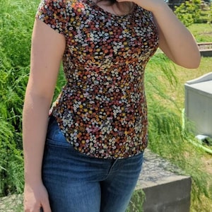 Puede incluir: Una blusa de manga corta con un denso estampado floral multicolor de pequeñas flores en tonos amarillo, blanco, naranja y rosa sobre un fondo negro, combinada con pantalones vaqueros de mezclilla azul oscuro y un sombrero de paja con cinta clara.