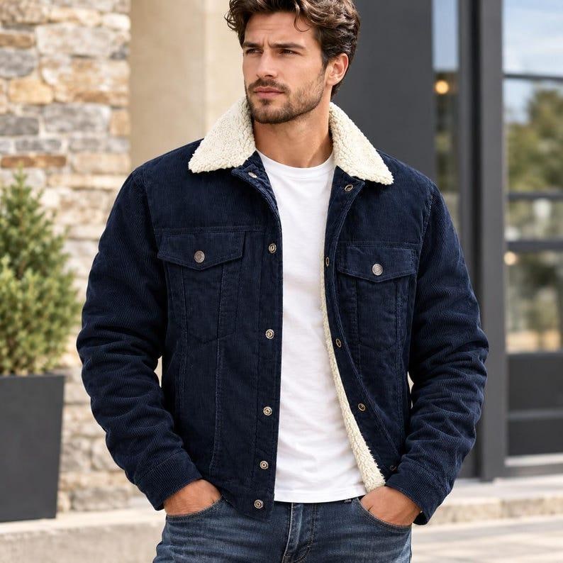 May include: A dark navy blue corduroy jacket featuring a white sherpa fleece collar and lining, a button-front closure, two chest pockets with buttoned flaps, and long sleeves. The jacket is worn over a plain white t-shirt and blue denim jeans.