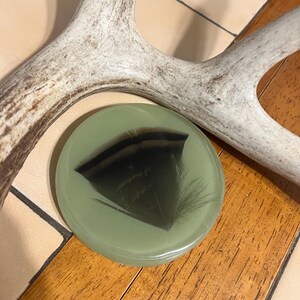 Op de afbeelding: Een ronde, saliegroene onderzetter of decoratieve schijf van hars met een enkele, donkergepunte veer in het midden. De veer heeft een opvallende zwarte band over de breedte en is ingebed in het gladde, ondoorzichtige groene oppervlak.