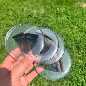 Op de afbeelding: Een set van drie ronde, transparante acryl onderzetters, elk met een donkerbruine en zwart gestreepte veer ingebed in het heldere materiaal. De onderzetters hebben gladde, afgeronde randen en een glanzende afwerking, waardoor het natuurlijke patroon van de veren duidelijk zichtbaar is door het transparante oppervlak.