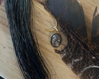 Op de afbeelding: Een goudkleurige slangenketting met een ovaalvormige hanger met een donkere, getextureerde, haarachtige inleg. De hanger is gevat in een dunne goudkleurige metalen zetting en hangt aan een fijne, gladde, goudkleurige ketting die op een oppervlak rust.