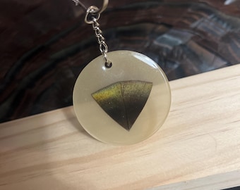 Op de afbeelding: Een ronde sleutelhanger van doorschijnende, gebroken witte hars met een subtiele glans. In het midden is een driehoekig, donkergekleurd veerfragment ingebed met een verloop van zwarte, bruine en iriserende goudtinten. Een zilverkleurige metalen ring en ketting zijn aan de bovenkant bevestigd.