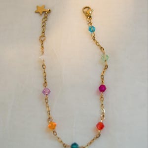 Puede incluir: Una pulsera de cadena en tono dorado con una serie de pequeñas cuentas facetadas multicolores en tonos azul, verde, rosa, naranja y rojo. La pulsera incluye un pequeño dije de estrella dorada en un extremo y un cierre de mosquetón.