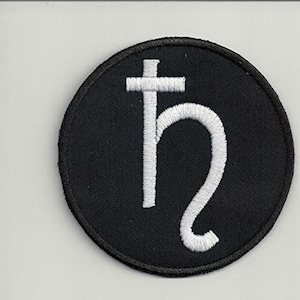 Pode incluir: Um remendo bordado preto e branco com o símbolo astrológico de Saturno, uma letra estilizada "h" com uma cruz no topo.