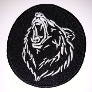 May include: A circular black embroidered patch with a white outline of a roaring bear's head. The bear's mouth is open, revealing its teeth. This patch is designed for application on clothing or accessories.
