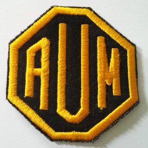 Puede incluir: Un parche negro en forma de octógono con un contorno amarillo y las letras "AUN" bordadas en amarillo.