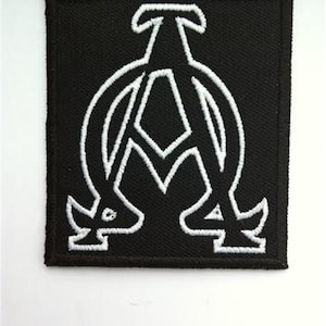 Puede incluir: Parche bordado negro con un contorno blanco de las letras griegas Alfa y Omega.