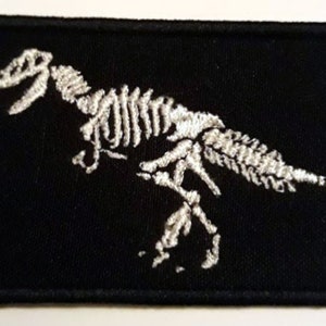 Puede incluir: Parche de tela negro con un esqueleto de dinosaurio bordado en blanco.