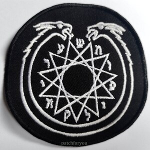Pode incluir: Patch bordado em preto e branco com um design circular. O círculo apresenta uma estrela branca de oito pontas com um centro preto e letras hebraicas ao redor da estrela. Duas cabeças de dragão brancas estão acima da estrela.
