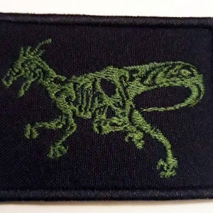 Puede incluir: Parche bordado verde sobre fondo negro con la silueta estilizada de un dinosaurio.