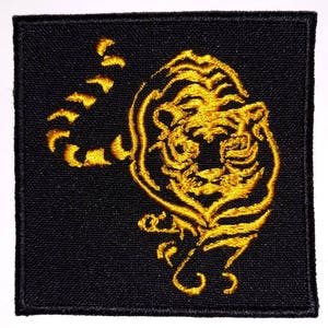 Op de afbeelding: Een gouden geborduurde patch met een tijger met zijn hoofd naar de zijkant gedraaid. De tijger is omrand met gouden draad op een zwarte stoffen achtergrond.