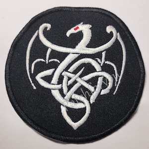 May include: Black embroidered patch featuring a white dragon design with a red eye and a Celtic knot. The patch has a rounded, oval shape and is made of fabric.