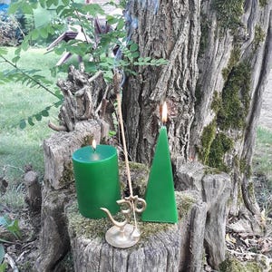 May include: A green cylindrical candle and a green triangular candle are lit on a mossy tree stump. An incense stick is placed in a gold-coloured incense holder with the Om symbol. The background includes a tree trunk and foliage.