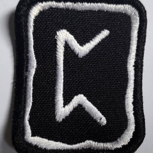 Pode incluir: Patch de tecido preto com um símbolo de runa bordado em branco e uma borda branca. A runa é chamada "Ehwaz" e representa a parceria, a viagem e o movimento.