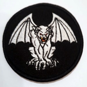 Puede incluir: Un parche bordado en blanco y negro de una gárgola con alas. La gárgola es blanca con ojos rojos y está sobre un fondo negro.