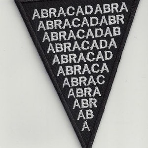 Pode incluir: Um remendo triangular preto com texto bordado branco que diz "ABRACADABRA" em um padrão descendente, começando com a palavra completa no topo e terminando com a letra "A" na parte inferior.