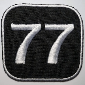 Puede incluir: Parche de tela negro con números bordados blancos "77" en forma cuadrada.