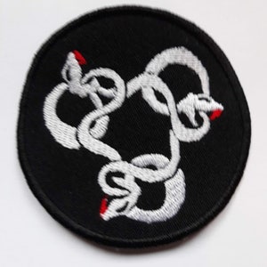 Pode incluir: Patch circular preto com um design de cobra branca bordada. A cobra está entrelaçada e tem acentos vermelhos na cauda.