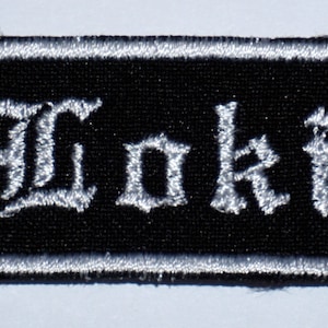 May include: Black embroidered patch with a white outline and the word "Loki" in white, stylized lettering.