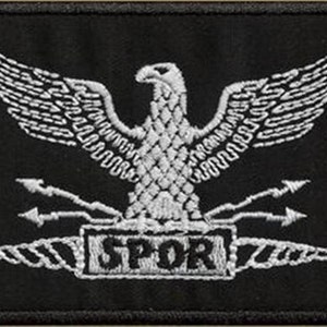 Op de afbeelding: Een wit geborduurde patch op een zwarte achtergrond met een Romeinse adelaar met uitgespreide vleugels, die een bundel pijlen in zijn klauwen houdt. De adelaar staat op een basis met het woord "SPQR" erop.
