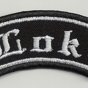 Può includere: Toppa ricamata in bianco e nero con la parola "Loki" in lettere bianche su sfondo nero. La toppa ha un bordo bianco ed è a forma di rettangolo curvo.