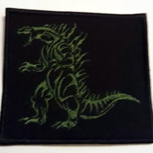 Puede incluir: Parche bordado verde de un dragón estilizado sobre un fondo negro.