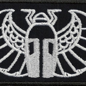 May include: White embroidered patch on a black background depicting a stylized winged scarab beetle.