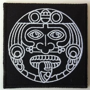 Peut inclure: Patch brodé noir et blanc représentant un visage stylisé avec des détails complexes. Le visage a de grands yeux, un nez proéminent et une large bouche. Le design rappelle l'art maya antique.