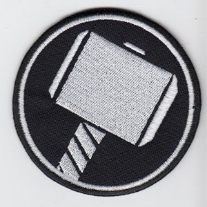 Könnte beinhalten: Ein gestickter Patch in Schwarz und Weiß mit einem stilisierten Hammer, der an Thors Hammer Mjolnir erinnert.