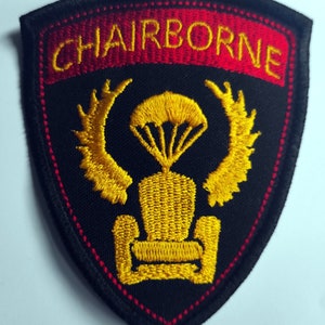 Könnte beinhalten: Ein schwarzer gestickter Aufnäher mit roten und goldenen Nähten. Der Aufnäher zeigt das Wort "AIRBORNE" in goldenen Buchstaben und einen stilisierten Fallschirm mit Flügeln.