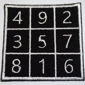 Könnte beinhalten: Ein schwarzes Stoffquadrat mit weißem Rand und weißen Zahlen 4, 9, 2, 3, 5, 7, 8, 1 und 6, die in einem 3x3-Raster angeordnet sind.
