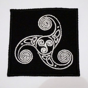 May include: Black square patch with a white embroidered Triskelion symbol. The Triskelion design features three spiraling arms connected by Celtic knotwork. The patch is likely for clothing or accessories.