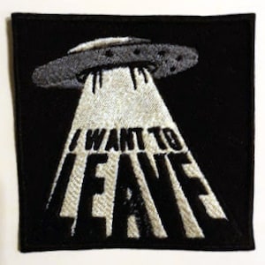 May include: Black square patch featuring a gray UFO emitting a white light beam. The text "I WANT TO LEAVE" is embroidered in bold, white letters within the beam. The patch has a black border.