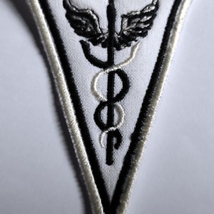Puede incluir: Parche triangular blanco con un símbolo médico bordado en negro, un caduceo con alas, sobre un fondo blanco con un borde blanco.