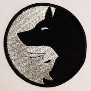 May include: A black and white embroidered patch with a wolf's head on one side and a cat's head on the other side.