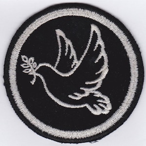 Op de afbeelding: Een geborduurde patch in zwart en wit met een witte duif die een olijftak in zijn bek houdt. De duif bevindt zich in een witte cirkel met een zwarte rand.