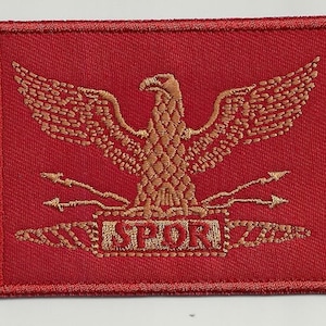 Può includere: Toppa ricamata con un'aquila dorata con ali spiegate, frecce e le lettere "SPQR" su sfondo rosso. La toppa è rettangolare con un bordo cucito.