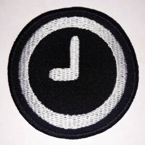Könnte beinhalten: Schwarz-weiß gestickter Aufnäher mit einem weißen Uhrzeiger in einem Kreis.