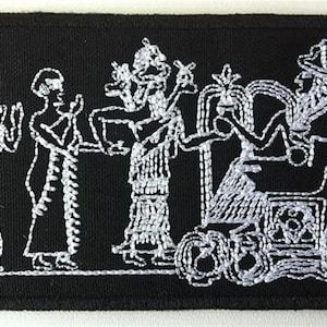 May include: White embroidered patch on a black background depicting five figures in a procession. The figures are stylized and appear to be wearing robes and headdresses. The figure in the center is seated on a throne with wheels.