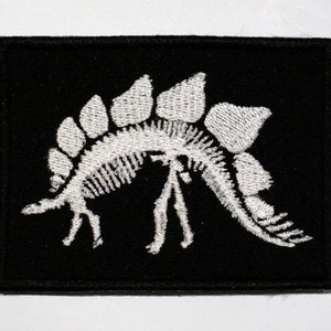 Puede incluir: Parche blanco bordado con un esqueleto de dinosaurio sobre un fondo de tela negro.