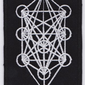 Op de afbeelding: Wit geborduurd geometrisch ontwerp op een zwarte stoffen patch. Het ontwerp is een complex patroon van elkaar kruisende lijnen en cirkels, dat lijkt op een levensboom.