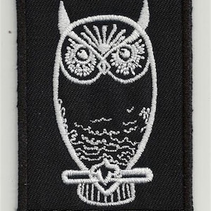 Peut inclure: Patch brodé blanc en forme de hibou sur fond noir. Le hibou est de face, les ailes repliées. Le patch est bordé de blanc.